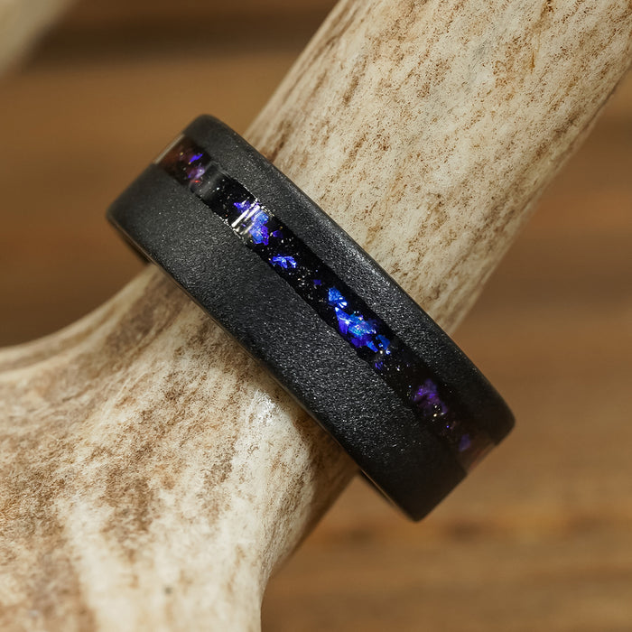 Introducing The Burro Creek: a striking black tungsten carbide ring featuring a central inlay of blue, purple, and iridescent flecks, displayed resting on a textured, light-colored antler. This bold men’s 8mm wedding band effortlessly combines durability with captivating modern design—perfect for the contemporary groom.