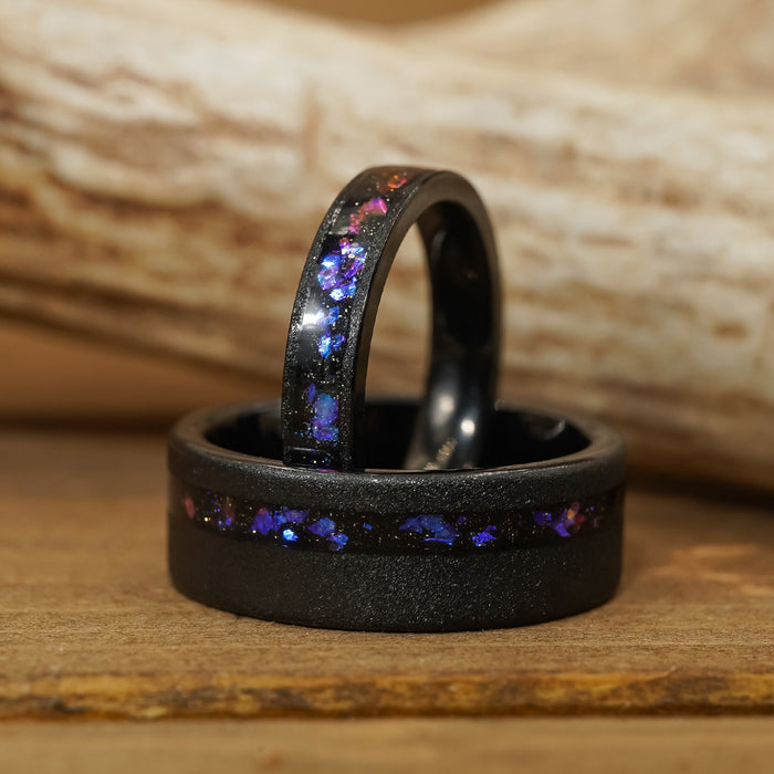 Two "Burro Creek" black tungsten carbide rings, inlaid with shimmering blue and purple fragments, are displayed on a wooden surface with a blurred log in the background—a striking choice for those seeking an 8mm men’s wedding band that combines modern style with unique detail.