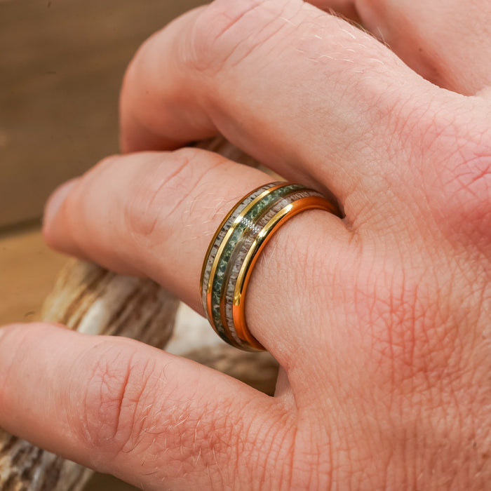 A close-up of a hand wearing The Butano, a gold tungsten carbide ring featuring two greenish inlays and shiny edges, resting on a piece of wood.