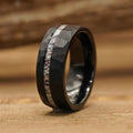 The Cades Cove, a black tungsten carbide ring with a faceted surface and a central inlay featuring a light, marbled pattern, is placed on a wooden surface.