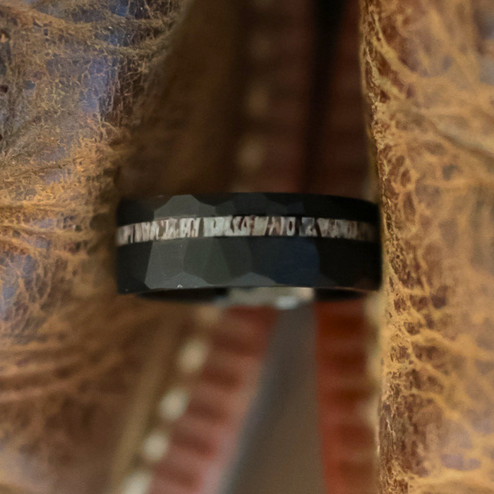 The Cades Cove, a black faceted tungsten carbide ring featuring a metallic inlay, is displayed between two pieces of worn brown leather.