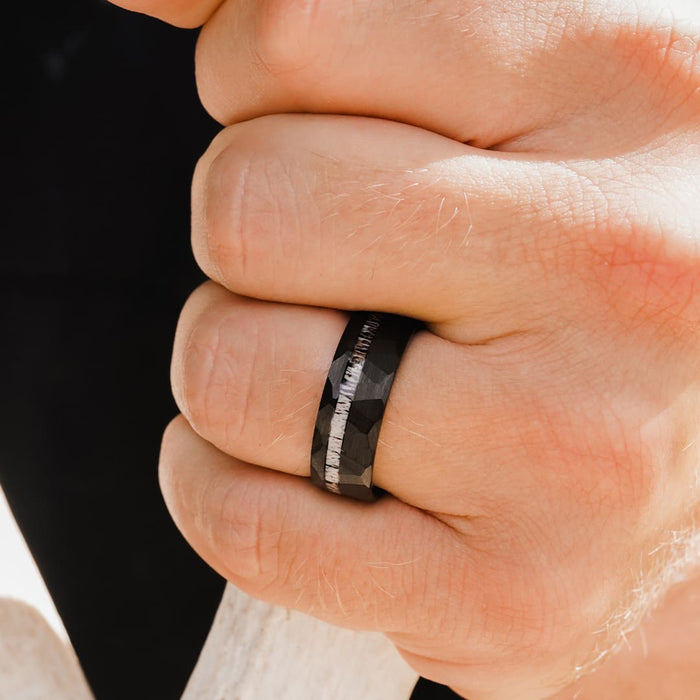 A close-up of a hand wearing The Cades Cove, a men’s hammered wedding band featuring a textured metallic stripe in the center. The hand is holding a light-colored object, with the background softly blurred.