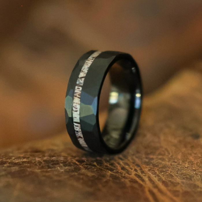 The Cades Cove, a black faceted tungsten carbide ring featuring a shiny patterned inlay, is displayed upright on a brown, textured surface.