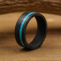 The Chesapeake Bay, an 8mm men’s ring crafted from forged carbon fiber with a striking crushed turquoise inlay, stands upright on a wooden surface, framed by a blurred, curved wooden background behind it.