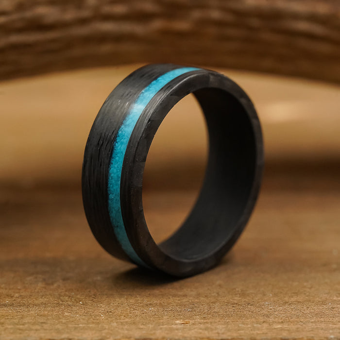 The Chesapeake Bay, an 8mm men’s ring crafted from forged carbon fiber with a striking crushed turquoise inlay, stands upright on a wooden surface, framed by a blurred, curved wooden background behind it.