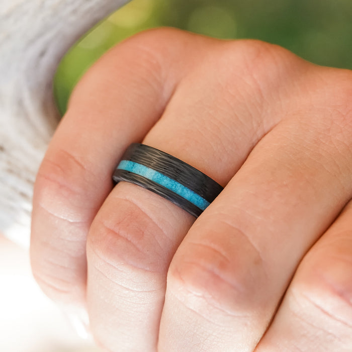 A hand wearing The Chesapeake Bay ring—an 8mm men’s band with a vibrant crushed turquoise inlay and forged carbon fiber—shown close-up against a blurred green background.