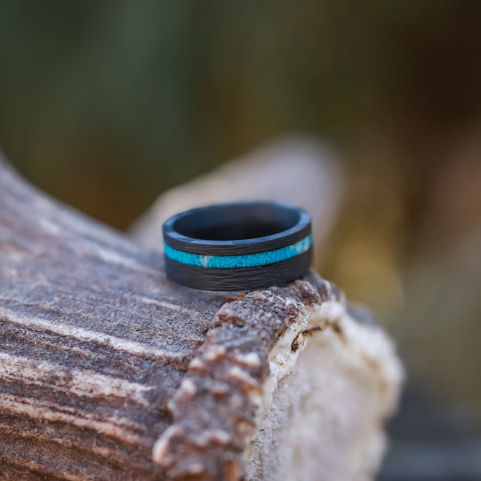 The Chesapeake Bay, a black men’s 8mm ring featuring a vibrant crushed turquoise inlay, rests atop a textured brown antler with a softly blurred natural background.