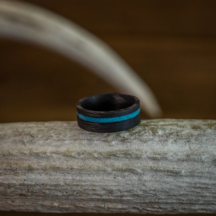 The Chesapeake Bay, a dark wooden ring featuring a crushed turquoise inlay, rests on a textured, light-colored branch set against a softly blurred brown background.