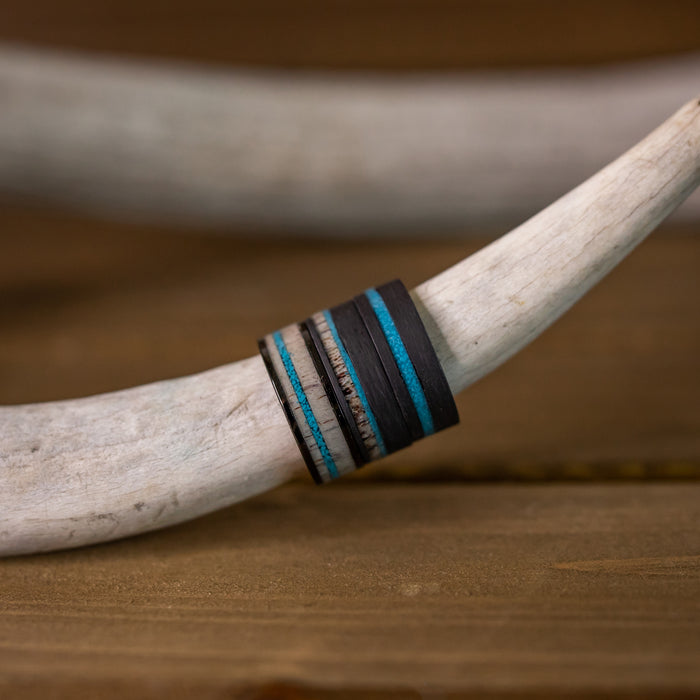 The Chesapeake Bay, an 8mm multi-layered men's ring featuring bands of black forged carbon fiber, brown and silver accents, and a bold crushed turquoise inlay, is displayed resting on a curved white antler against a wooden background.