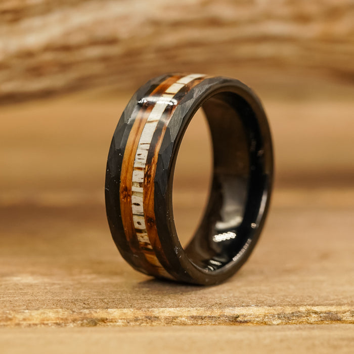 Displayed on a wooden surface against a softly blurred natural backdrop, The Custer is a striking black ring with a polished finish. Its design features inlaid wood and a metallic strip, evoking the craftsmanship of whiskey barrel rings and blending refined elegance with rustic charm.