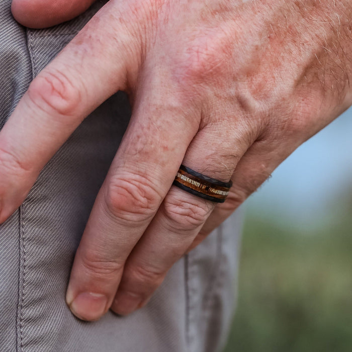 A close-up of a person's left hand resting on their pants, showcasing The Custer—a striking hammered tungsten ring—on their ring finger. The hand appears relaxed against a blurred background.