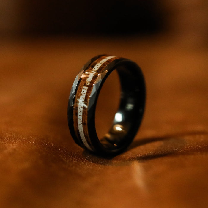 A close-up of The Custer, a black hammered tungsten ring featuring two distinct inlaid stripes—one brown, inspired by whiskey barrel wood, and one silver—resting on a soft brown surface.