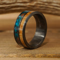 The Desoto, a black brushed ring featuring both a wood inlay and a turquoise stone inlay, is displayed upright on a wooden surface against a softly blurred wooden background.