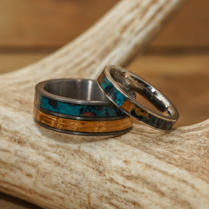 Two metal rings rest on a piece of antler. One is The Desoto, a wide tungsten carbide ring featuring striking blue and gold bands, while the other is a slimmer black brushed ring adorned with a silver and dark patterned design. The background is blurred wood.