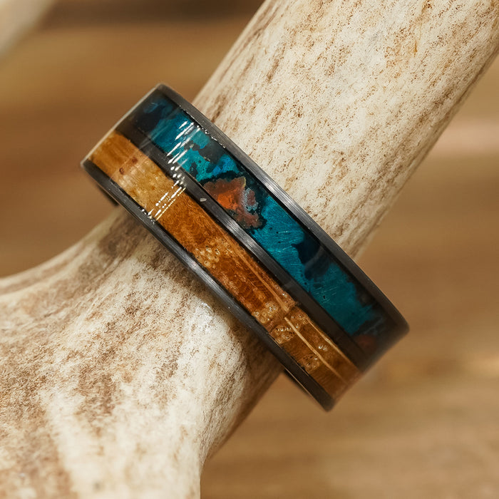 A close-up of The Desoto ring, featuring a black brushed metal band with a wood inlay and turquoise-colored resin accents, resting on a beige antler. The ring showcases a dark finish and glossy detailing.