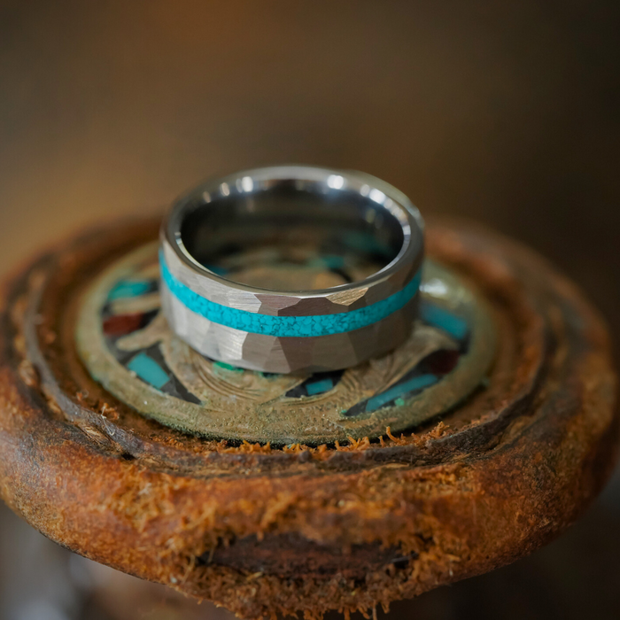 The Dry Tortuga, a hammered tungsten ring featuring a crushed turquoise inlay and camouflage pattern, rests on a rustic circular surface adorned with turquoise and brown accents.