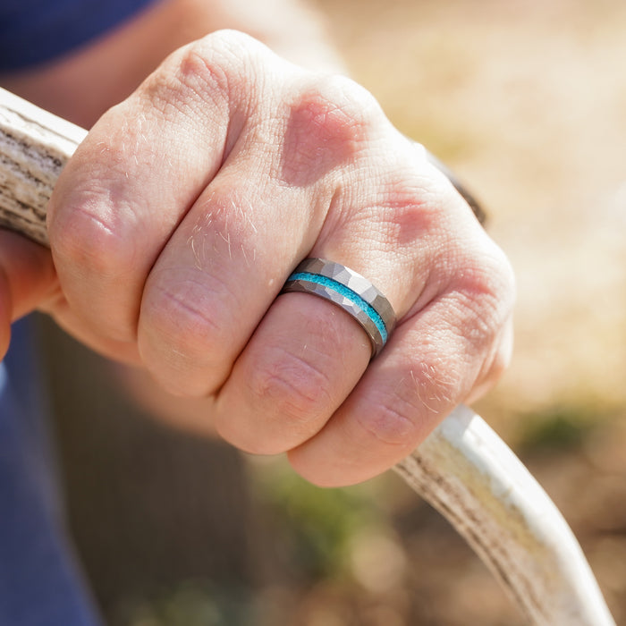 A close-up of a hand gripping a white antler showcases The Dry Tortuga, a hammered tungsten ring with a blue crushed turquoise inlay, worn on the person's ring finger. The background is slightly blurred.