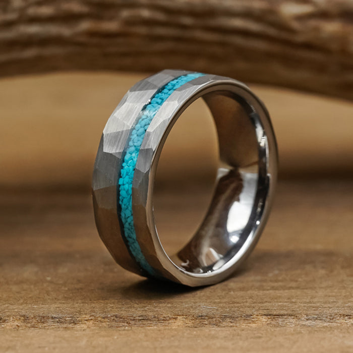 Introducing The Dry Tortuga: a silver-toned ring featuring a faceted, hammered tungsten surface and a slim inlay of vibrant crushed turquoise stones encircling the center, elegantly resting on a wooden surface.