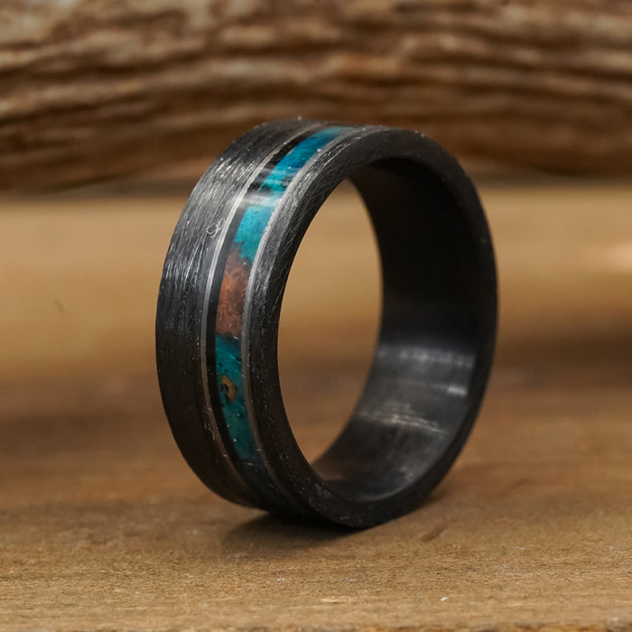 The Eagle Canyon ring, crafted from durable Tungsten Carbide with an 8mm width, showcases a striking black brushed finish and a turquoise and brown inlay stripe. It stands upright on a wooden surface, set against a softly blurred wood background—blending modern elegance with rugged resilience.