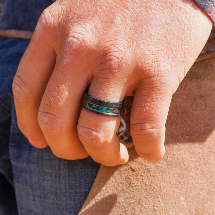 A close-up of a hand wearing The Eagle Canyon, an 8mm wide black brushed Tungsten Carbide ring featuring a blue inlay, resting on a knee. The person is dressed in blue jeans and a shirt with rolled sleeves.