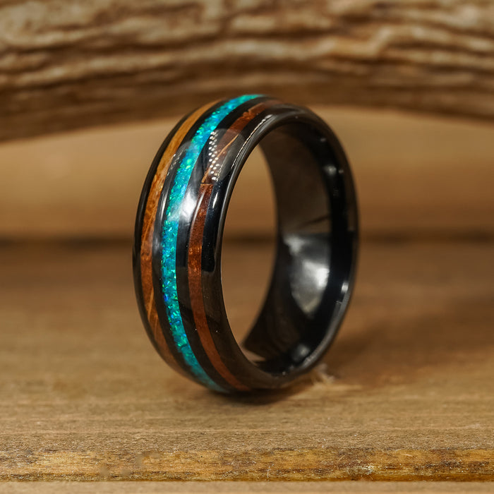 The El Capitan, a black tungsten carbide ring featuring a shiny turquoise inlay and a wooden stripe, sits on a wooden surface against a blurred wood-grain background.