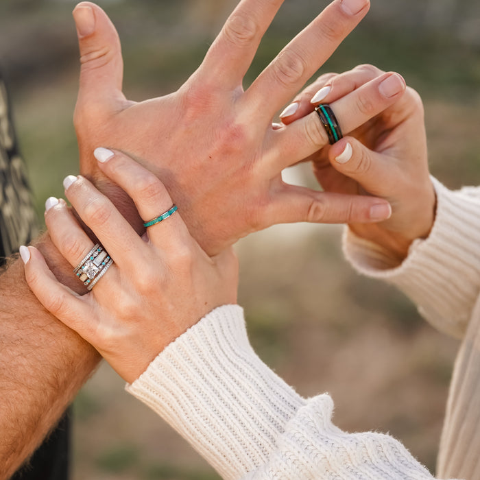 Two hands, each adorned with rings, meet in a gentle touch. One hand is wrapped in a white sweater sleeve with polished nails, while the other remains bare. Amidst the turquoise and silver bands on their fingers, The El Capitan ring—with its opal inlay—shimmers with iridescent hues as it catches the light.
