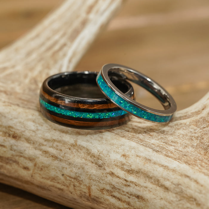 Two El Capitan rings with turquoise inlays rest on a tan antler. The wider El Capitan ring showcases a striking wood accent reminiscent of a whiskey barrel, while the slimmer band is crafted entirely from metal. The background features softly blurred natural wood tones.