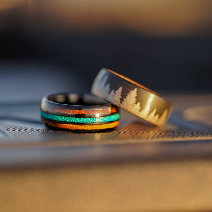 Two rings rest side by side: The El Capitan, an opal inlay ring featuring blue-green shimmer and wood accents, and a matte black tungsten carbide ring engraved with pine trees. Both are displayed on a textured surface, softly lit by warm sunlight.