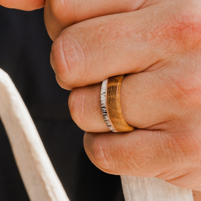 A close-up of a hand wearing The Faroe, a brown and white Whiskey Barrel Ring featuring a natural, wood-like texture and metallic accents that showcase its unique design and craftsmanship.