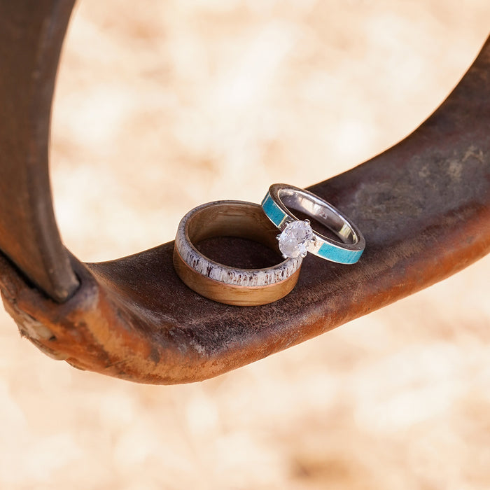 Two rings rest on a rusty metal surface: one is The Faroe, a silver band with turquoise inlay and a diamond solitaire, while the other is a thicker, rustic Whiskey Barrel Ring crafted from silver and wood. The background is blurred.