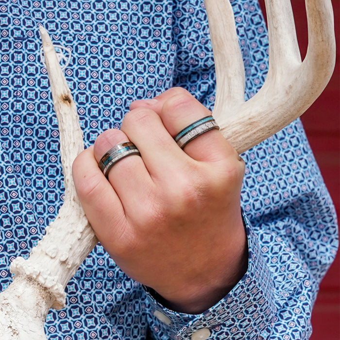 A person wearing a blue patterned shirt holds a white deer antler, with two silver and blue rings—one being the striking "The Folsom Lake" tungsten carbide ring—visible on their fingers. The background is red.