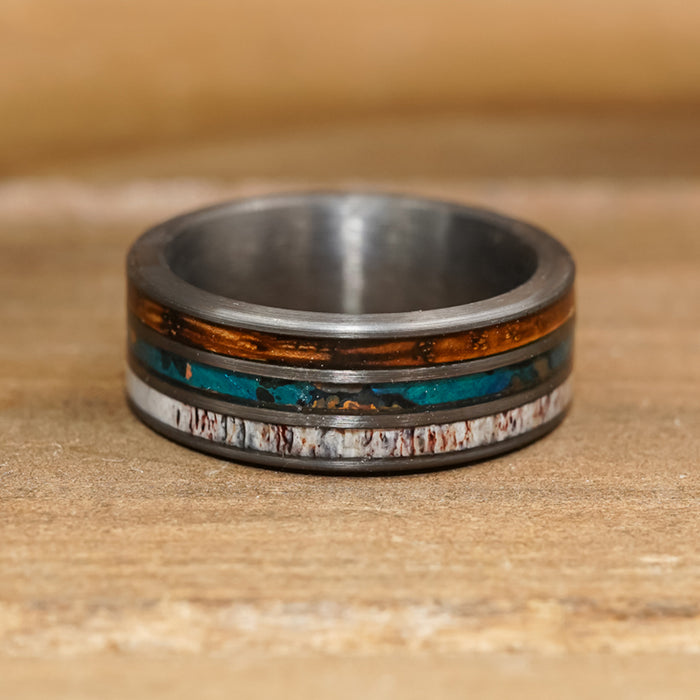 The Folsom Lake ring, crafted from tungsten carbide, features three distinctive inlay bands—a top amber-brown strip, a middle blue-green strip, and a bottom white strip with dark marbling—displayed on a wooden surface.