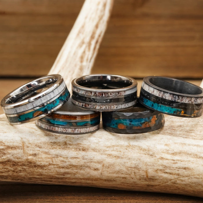 Five metallic rings, featuring "The Folsom Lake"—a striking tungsten carbide ring with turquoise and brown inlays—alongside a patina copper ring, rest on a light-colored antler against a wooden background. The rings showcase rustic, unique designs that echo the natural appeal of an antler ring.