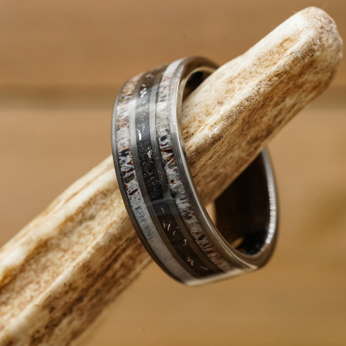 The Fossil Ridge, a black and silver tungsten carbide ring featuring a textured, multicolored inlay, is displayed on a light brown, textured bone against a blurred wooden background.