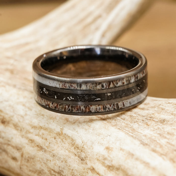 A close-up of The Fossil Ridge, a dark metal tungsten carbide ring with lighter, textured inlays, rests on a light brown antler.