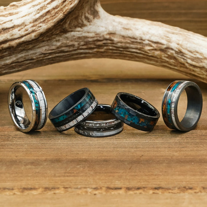 Five unique men's rings, including The Fossil Ridge with its distinctive design, feature various inlays such as turquoise, wood, and a striking meteorite ring. They are displayed on a wooden surface in front of a large antler.