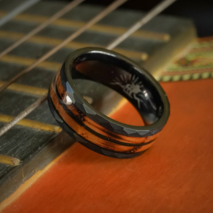 The Gatlinburg, a black and wood-grain tungsten carbide ring with a faceted edge, rests on the strings and fretboard of an acoustic guitar.