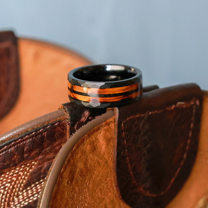 A glossy-finished "The Gatlinburg" whiskey barrel ring, featuring black and brown stripes, rests on the folded edge of a brown leather item—possibly a wallet or bag—set against a light blue background.