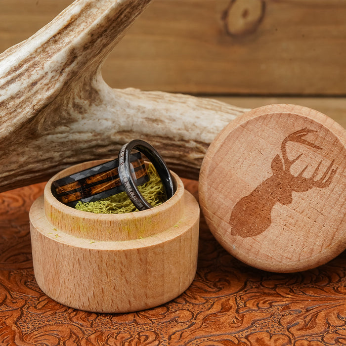 The Gatlinburg—featuring a tungsten carbide ring and a whiskey barrel ring—rests in a small round wooden box filled with moss. The nearby lid showcases an engraved silhouette of a buck, while an antler and tooled leather provide the perfect rustic backdrop.