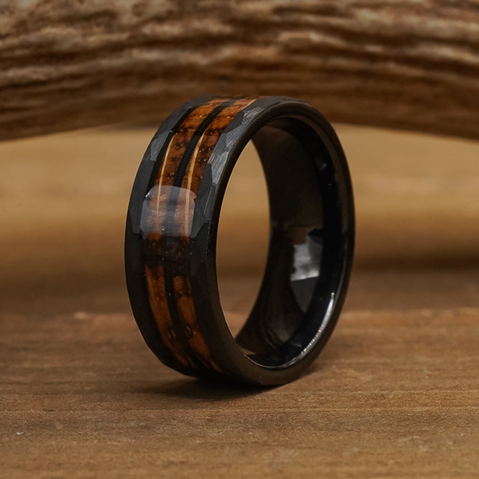The Gatlinburg, a black tungsten carbide ring featuring two parallel brown wood inlay stripes reminiscent of a whiskey barrel, rests on a wooden surface with a wooden object softly blurred in the background.