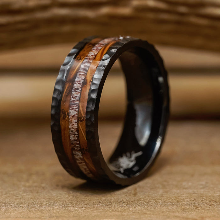 The Gifford, a black men's hammered ring featuring a wood inlay and copper stripe, is displayed on a wooden surface with a softly blurred wooden background.