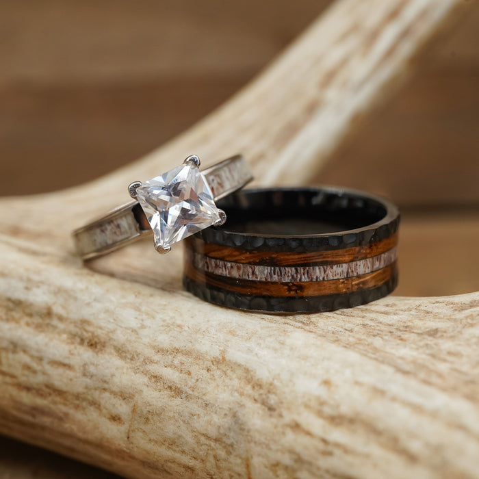 Two rings rest on a piece of antler: one is The Gifford, an elegant antler ring with a square-cut clear stone, while the other is a black titanium wedding band featuring striking wood and antler inlays.