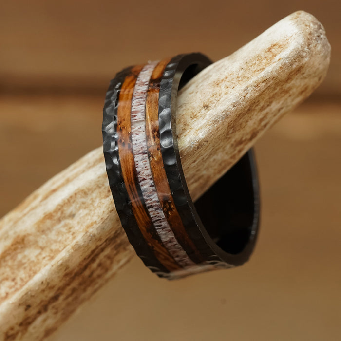 The Gifford is a black metal antler ring with a hammered texture, showcasing two wooden inlays and a light-colored central stripe, elegantly displayed on an antler.