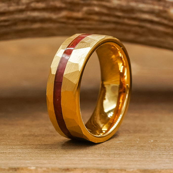The Golden Canyon ring, featuring a faceted gold-plated surface and a single red inlay band, stands upright on a wooden surface with a blurred piece of wood in the background.