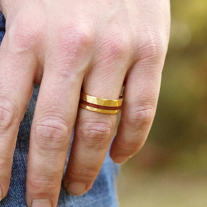 A close-up of a person’s hand wearing two hammered gold rings, including The Golden Canyon gold plated ring, on the ring finger. The hand is relaxed and partially clenched, with a blurred background.