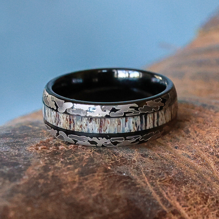 The Goliad, a black tungsten carbide ring featuring a central band of textured, multicolored metal with engraved camouflage patterns, rests on a brown textured surface against a blue background—making it a striking choice for a men’s wedding band.