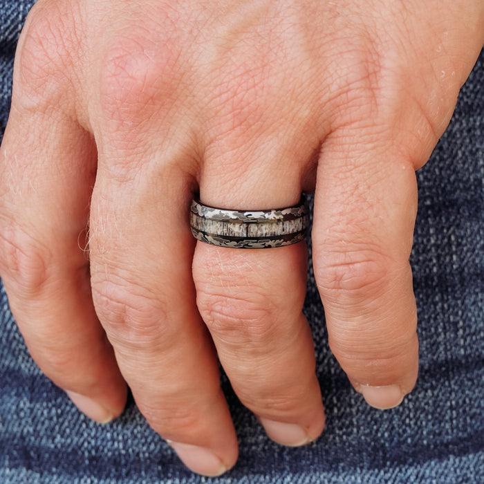 A close-up of a hand wearing The Goliad, a gray patterned tungsten carbide ring, on the middle finger, with blue denim fabric in the background.
