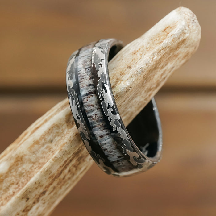 The Goliad is a striking silver and dark tungsten carbide ring featuring a leaf or camo pattern and a textured inlay. Uniquely crafted with an antler design, this ring is showcased elegantly on a piece of antler against a softly blurred brown background—making it the perfect wedding band for men who appreciate rugged sophistication.