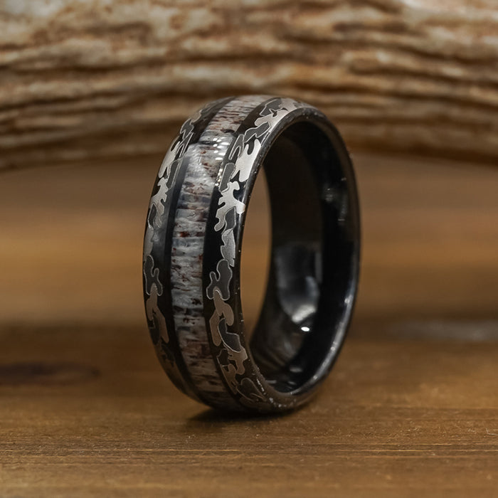 The Goliad, a black tungsten carbide ring with a camo-patterned inlay and wood-like texture, rests on a wooden surface against a blurred wooden background—making it the ideal wedding band for men seeking a rugged yet stylish accessory.