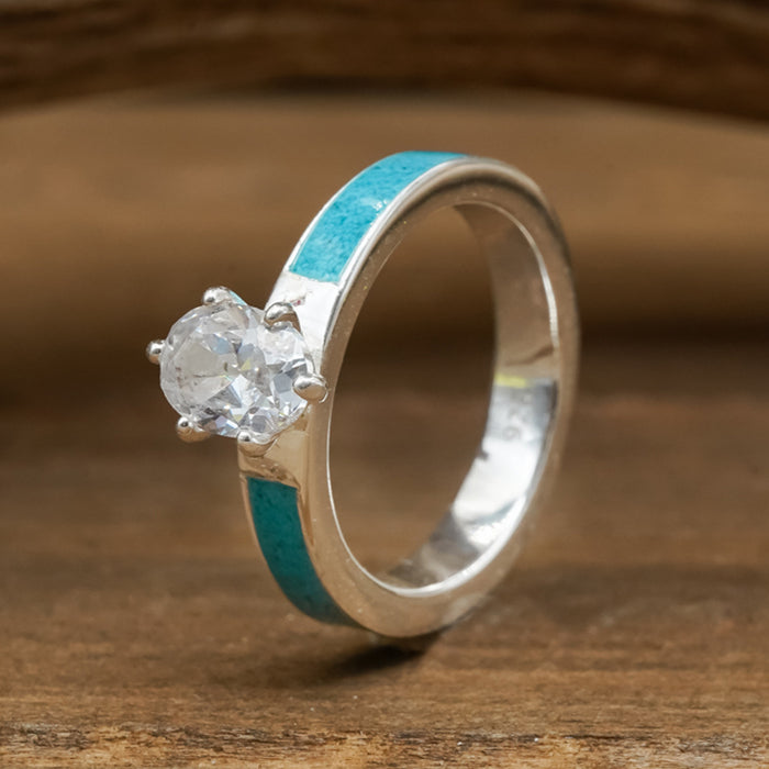 The Grand Adair - Turquoise Inlay is a sterling silver engagement ring showcasing a round, clear gemstone at its center and a vivid crushed turquoise inlay along the band, elegantly presented on a wooden surface.